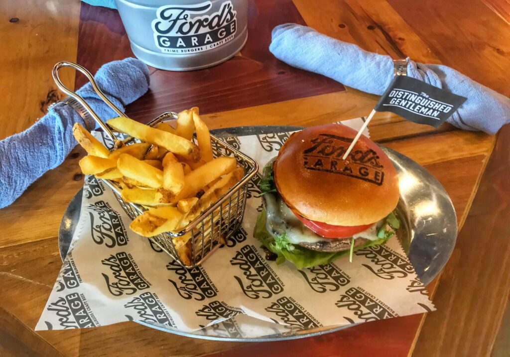 Image of a burger with a toothpick inserted in it that says "The Distinguished Gentleman", next to a basket of golden fries, from Ford's Garage.